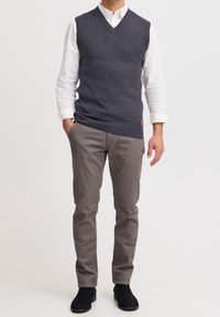 Pull en tricot sans manches gris foncé avec un col en V, porté sur une chemise blanche à boutons, associé à un pantalon gris clair et des chaussures noires.