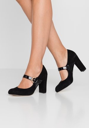 Musta mokkanahka korkokengät pyöreällä kärjellä, paksulla korolla ja kiiltävästä nahasta valmistettu nilkkaremmi, jossa on pyöreä solki.