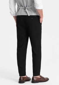 Pantaloni neri con un taglio su misura, caratterizzati da un tessuto liscio, orli risvoltati e una tasca posteriore discreta. Abbinati a scarpe marroni.