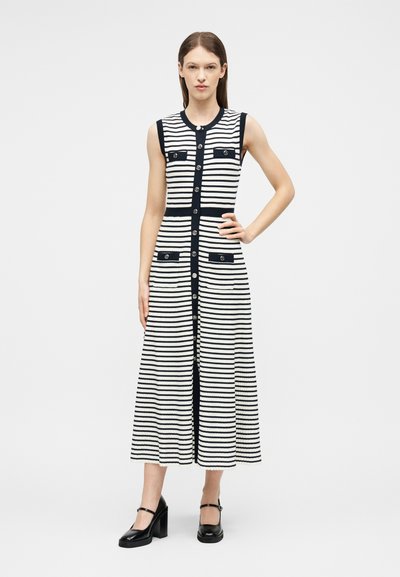 Femme debout, main sur la hanche, portant une robe midi sans manches à rayures noires et blanches avec des boutons sur le devant et des escarpins Mary Jane noirs.