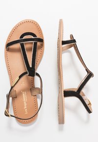 Black suede sandals with a minimalist design, featuring narrow straps and tan leather soles. Gold metal buckle detail on the ankle strap.