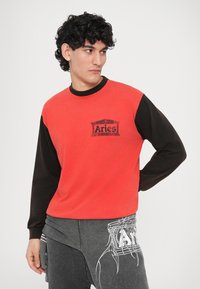 Duga crvena i crna majica dugih rukava s okruglim izrezom i tiskanim logotipom sprijeda. Meki materijal, ležerni kroj.