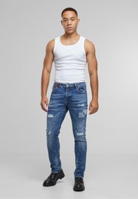 Jeans de mezclilla azul con un corte slim, que presentan parches desgastados, desvanecidos y manchas de pintura. El modelo lleva una camiseta sin mangas blanca y zapatos negros.