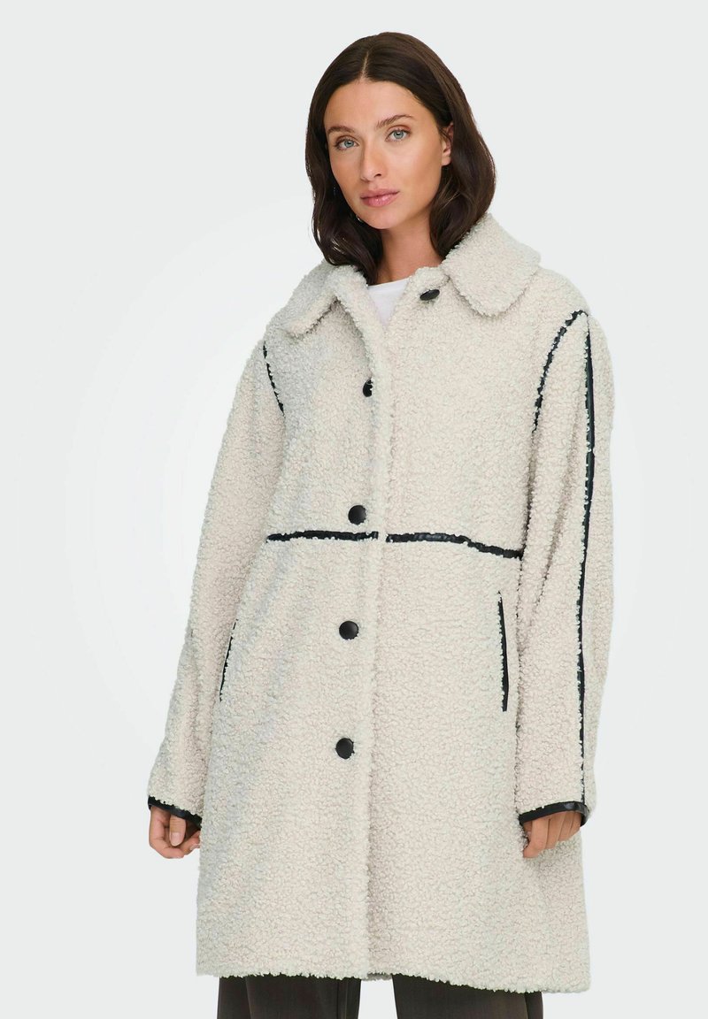 Cappotto in pile color crema con accenti neri, grande colletto, bottoni frontali e tasche laterali. Presenta una vestibilità morbida e comoda e una superficie testurizzata.