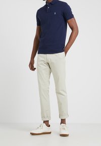 Granatowa koszulka polo z krótkim rękawem, z haftowanym logo na piersi, w zestawieniu z podwiniętymi jasnobeżowymi chinosami i białymi trampkami z brązowymi podeszwami.