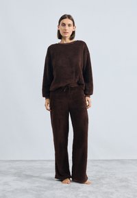 Brūna, mīksta mājas apģērba komplekts, izgatavots no pūkaina auduma; satur brīvi pieguļošu, garām piedurknēm topu un atbilstošas platās bikses ar elastīgu jostu.