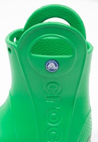 Zuecos de goma verde con textura de panal, parte trasera elevada, logo circular y detalle del logo en el lateral. Cuenta con una asa para facilitar su uso.