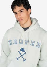 Joven con cabello corto oscuro que lleva una sudadera con capucha de color claro con el texto "HARPER" y un diseño de bates de cricket cruzados y una gorra.