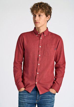 Camisa roja de botones con una tela texturizada y un bolsillo en el frente. Presenta un cuello clásico y botones blancos; combinada con jeans de mezclilla.