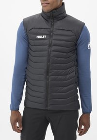 Gilet rembourré noir avec un col haut, présentant des coutures horizontales pour l'isolation et une fermeture éclair à l'avant. Porté par-dessus une chemise à manches longues texturée bleue.