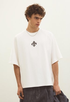 Mladý muž s kučeravými vlasmi nosí veľké biele tričko s čiernym dizajnom fleur-de-lis, tmavé džínsy, strieborný reťaz a drží rifľovú bundu.