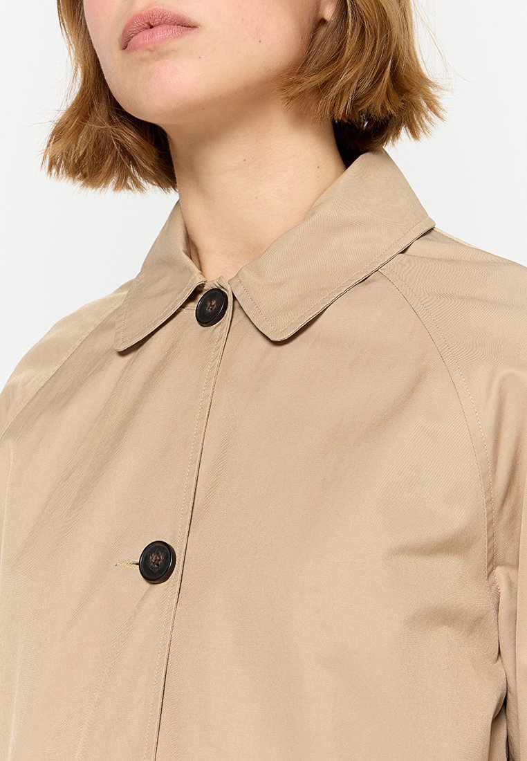 Cappotto beige con colletto classico, due grandi bottoni marroni e una texture liscia. Presenta maniche raglan e dettagli minimi.