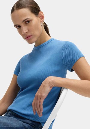 Žena s uhlazenými vlasy staženými dozadu, nosící kruhové náušnice a modrý texturovaný krátký top s rukávy, sedí a opírá ruku o bílou židli.