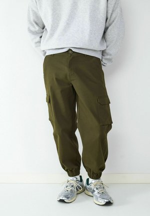 Person iført olivengrønne cargobukser med elastiske manchetter, lysegrå sweatshirt og sølvfarvede løbesko står op ad en hvid væg.