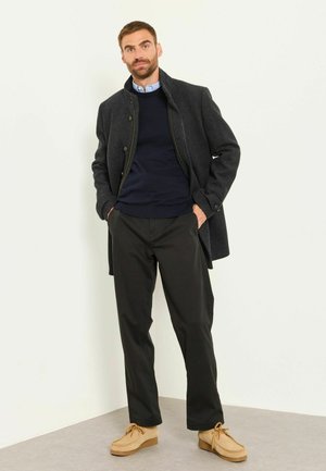 Mann trägt dunkelgrauen Mantel, marineblauen Pullover über hellblauem Hemd, schwarze Hose und beige Schuhe, steht mit den Händen in den Taschen.