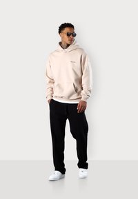 Beige Kapuzenpullover mit einer Fronttasche und gesticktem Logo, kombiniert mit schwarzen Trainingshosen und weißen Turnschuhen, stilvoll ergänzt durch dunkle Sonnenbrillen.