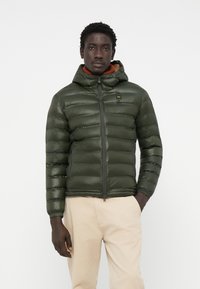 Padded green jacket with a zippered front and attached hood, featuring a smooth texture and elastic cuffs, paired with light trousers.