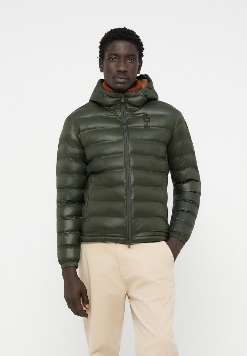 Padded green jacket with a zippered front and attached hood, featuring a smooth texture and elastic cuffs, paired with light trousers.