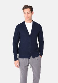 Cardigan navy lavorato a maglia con scollo a V, trama a coste, sei bottoni sul davanti e maniche lunghe. Indossato sopra una maglietta bianca e pantaloni grigi.