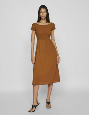 Robe brune mi-longue avec un corsage smocké et des manches courtes, jupe fluide et texture douce ; associée à des talons noirs à lanières.