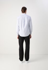 Camisa de botones a rayas azul claro con mangas largas, dobladillo redondeado y jeans negros. Parte de la espalda del modelo visible con zapatillas blancas en los pies.