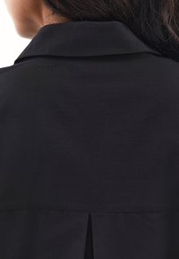 Nahaufnahme des oberen Rückens einer Person, die ein schwarzes kragenloses Kleidungsstück trägt, mit teilweise sichtbarem dunklem Haar.