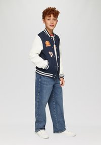 Chaqueta varsity azul marino con mangas blancas, que presenta parches y un cuello acanalado. Combinada con jeans azules holgados y zapatillas blancas.