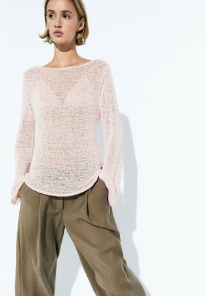 Junge Frau mit kurzen Haaren, die einen hellrosa locker gestrickten Pullover und lockere olivgrüne Hosen vor einem schlichten hellen Hintergrund trägt.