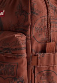 Sac à dos en tissu marron avec une surface texturée et des motifs imprimés noirs, y compris des logos et des illustrations. Détail de la fermeture éclair et de la poche latérale visible.