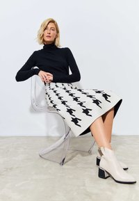 Tricou negru cu guler, fustă până la genunchi cu model houndstooth negru, botine bej cu tocuri groase, așezată pe un scaun din acrilic transparent.