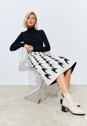 Tricou negru cu guler, fustă până la genunchi cu model houndstooth negru, botine bej cu tocuri groase, așezată pe un scaun din acrilic transparent.