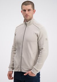 Beżowa kurtka na zamek z fakturowaną tkaniną na rękawach, bocznych kieszeniach i stójce. Posiada ściągacze na mankietach oraz subtelne logo.
