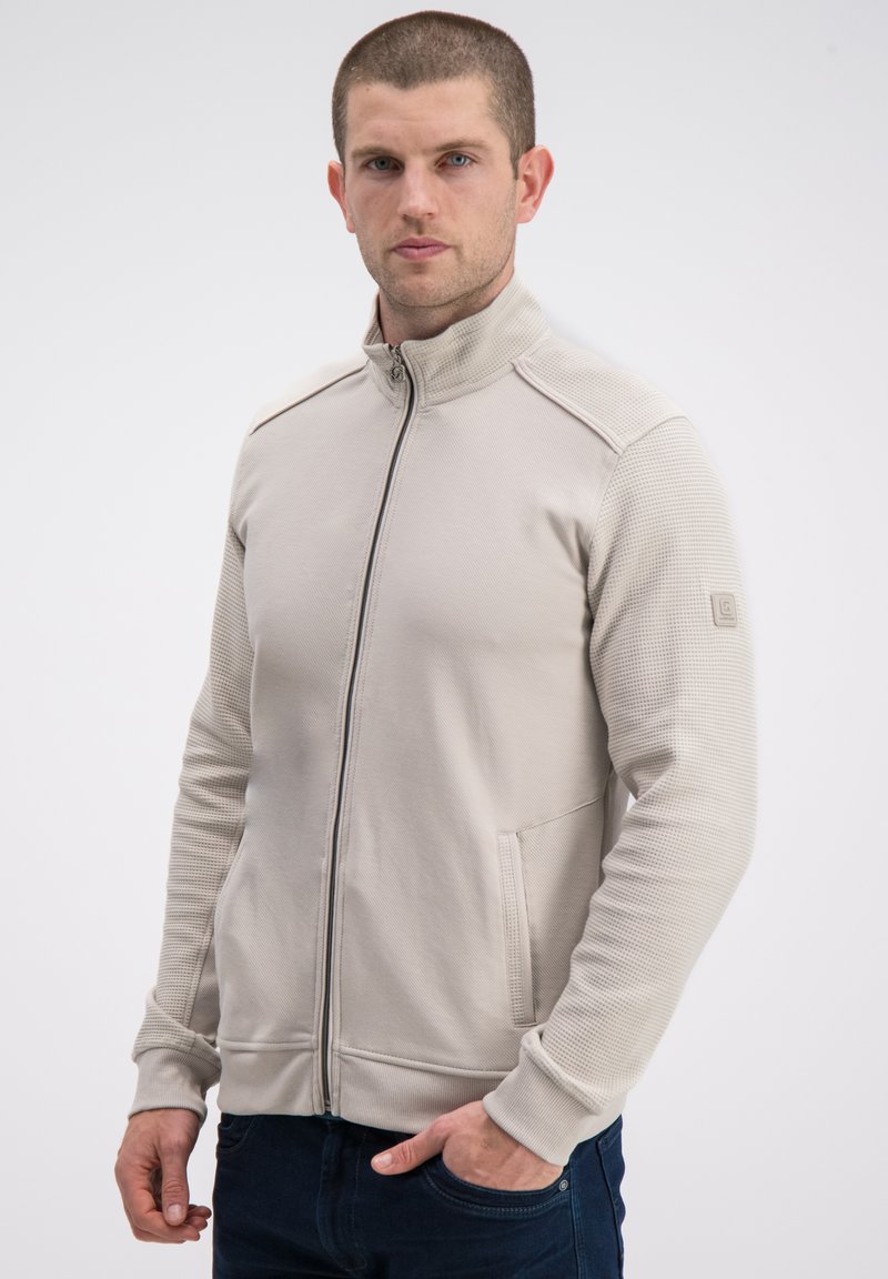 Beżowa kurtka na zamek z fakturowaną tkaniną na rękawach, bocznych kieszeniach i stójce. Posiada ściągacze na mankietach oraz subtelne logo.