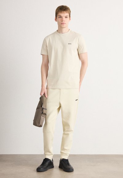 T-shirt en coton beige avec un petit logo noir "BOSS", associé à un jogging beige assorti et des baskets noires, tenant un sac marron.