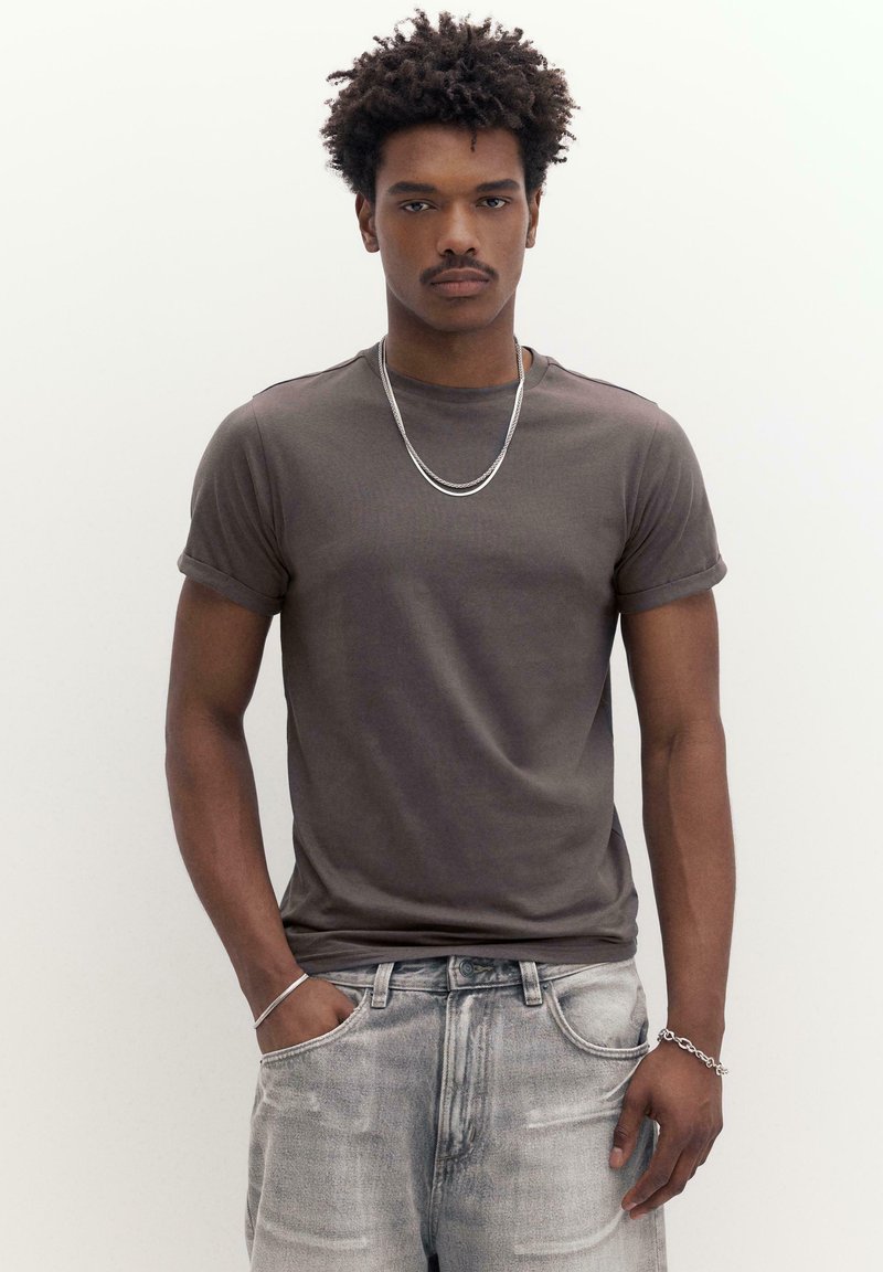 Jeune homme aux cheveux bouclés portant un t-shirt gris foncé à manches retroussées, un jean délavé, ainsi qu'un collier et des bracelets en chaîne argentés, debout devant un fond blanc.