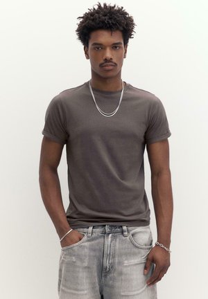 Giovane uomo con capelli ricci che indossa una t-shirt grigio scuro con maniche arrotolate, jeans sbiaditi e una collana e braccialetti d'argento, in piedi contro uno sfondo bianco.