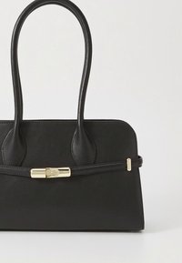 Svart strukturert håndveske med to avrundede håndtak og en slank rem med en lås i gulltone på hvit bakgrunn.