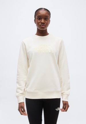 Vrouw draagt een crème kleurige trui en zwarte legging, beide met het logo van The North Face, staand tegen een effen lichte achtergrond.