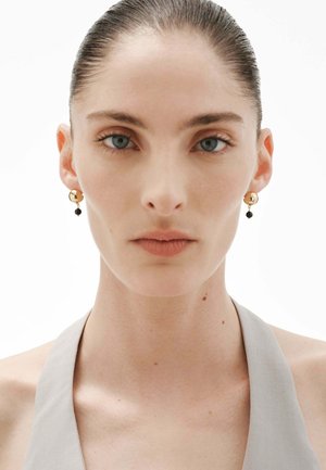 Mujer con el cabello hacia atrás, llevando pendientes de bolas de oro con cuentas negras y una blusa de halter gris claro, expresión facial neutra, fondo blanco.