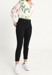 Blouse fleurie à manches longues, base blanche avec fleurs roses et vertes ; jean noir ajusté, tissu élastique, longueur cheville ; baskets blanches.