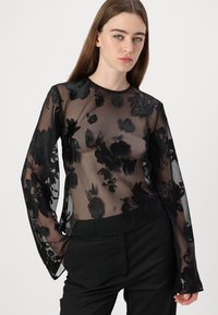 Gjennomsiktig svart topp med lange ermer, som har blomsterdesign. Glatt tekstur og rund halslinning, kombinert med skreddersydde svarte bukser.