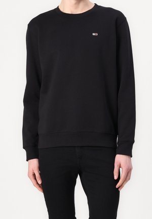 Homme portant un sweat-shirt noir uni à manches longues avec un petit logo rectangulaire rouge, blanc et bleu sur la poitrine gauche, associé à un pantalon noir.