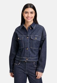 Tmavomodrá denimada bunda so strednými gombíkmi, dvoma prednými vreckami a kontrastným prešívaním. Krátky strih s golierom a uvoľneným strihom.