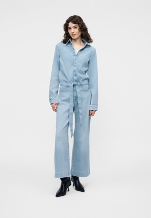 Žena s kučeravými vlasmi, oblečená v svetlomodrom rifľovom overale s opaskom a čiernymi členkovými čižmami na podpätku, stojaca na jednoduchom pozadí.