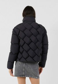 Persoon in een zwart geplooid pufferjack met grote vierkante stiksels en een hoge kraag, gecombineerd met een vervaagde grijze spijkerrok.