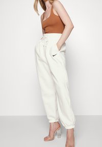 Žena oblečená v bílých Nike teplácích s šňůrkou, hnědém bezrukávovém topu a průhledných sandálech na vysokém podpatku, s rukama v kapsách, stojící na šedé podlaze.