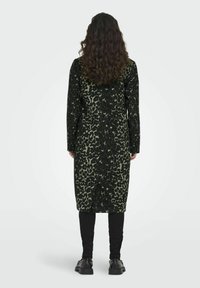 Cappotto lungo leopardato in nero e verde con maniche lunghe, tessuto testurizzato e taglio dritto, abbinato a pantaloni slim neri e scarpe chunky.