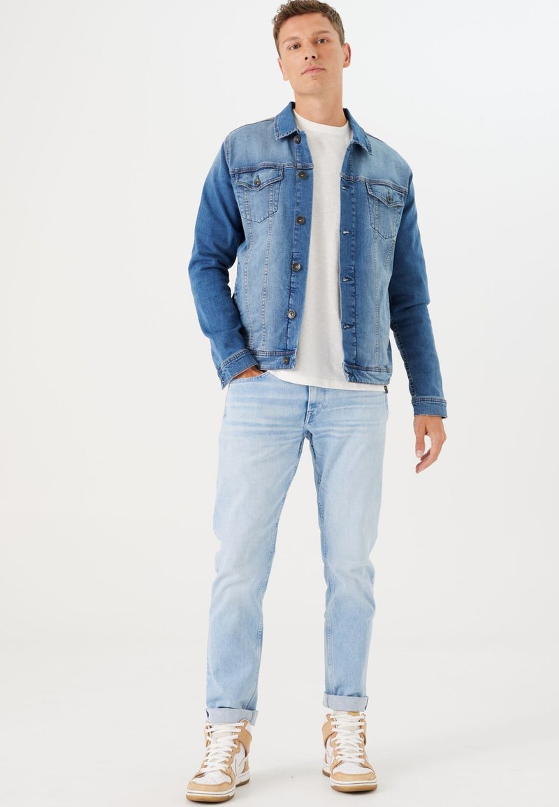 Chaqueta de denim con un degradado de azul claro y azul oscuro, con frente abotonado, dos bolsillos en el pecho y un corte relajado, combinada con jeans claros.
