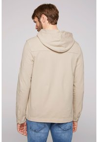 Chaqueta con capucha beige de textura suave, diseñada con mangas largas y un ajuste holgado, combinada con jeans de mezclilla azul claro.