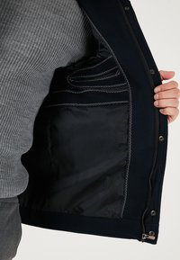 Námornícka vonkajšia bunda s hladkou čiernou podšívkou. Obsahuje podrobné prešívanie, zips a bočné vrecká. Nosená cez šedý pletený sveter.